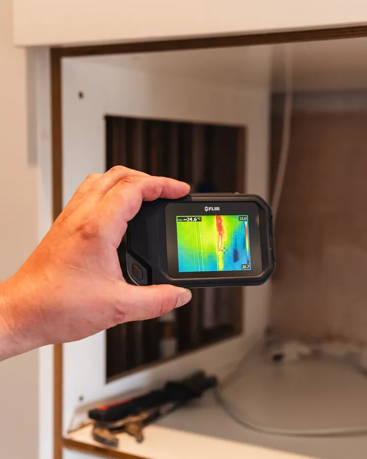 An ADI engineer holding a thermal camera towards concealed pipework, the screen showing a warm line running down the wall